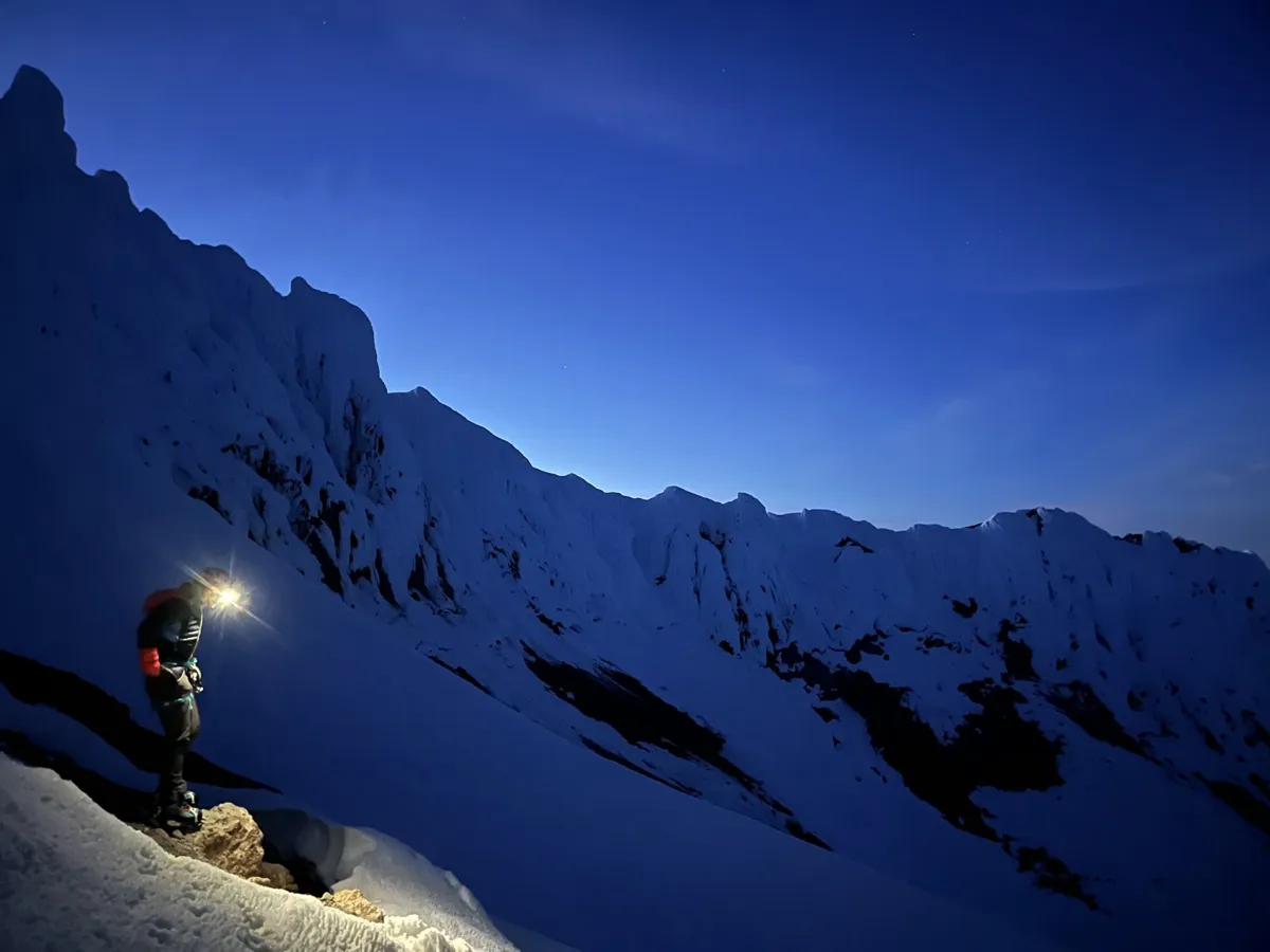 A climber with a headlamp on a snowy ridge below Mount Hood's cliffs before sunrise